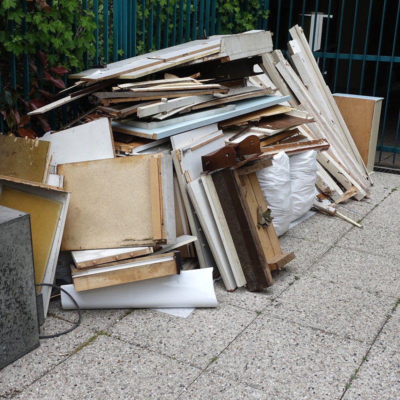 Grand tas de débris de bois, panneaux, vieux meubles et encombrants près d'une clôture verte sur un trottoir carrelé. Sacs blancs visibles.