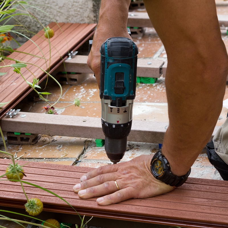 Main utilisant une perceuse pour fixer une lame de terrasse en bois composite. Pose du platelage sur lambourdes et plots.