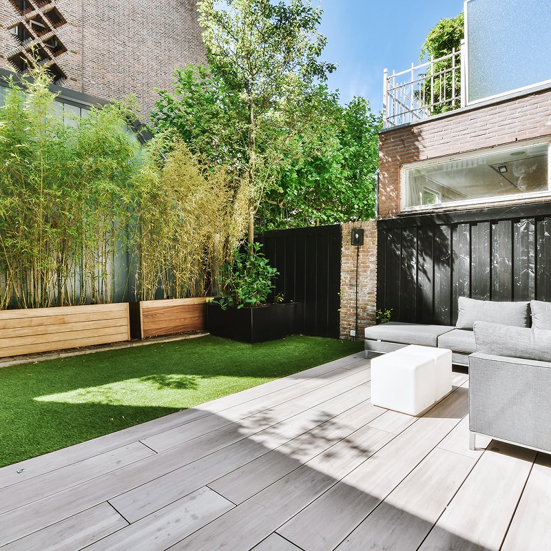 Terrasse moderne avec salon extérieur gris sur deck en bois, gazon synthétique, bambous en jardinières et mur de briques