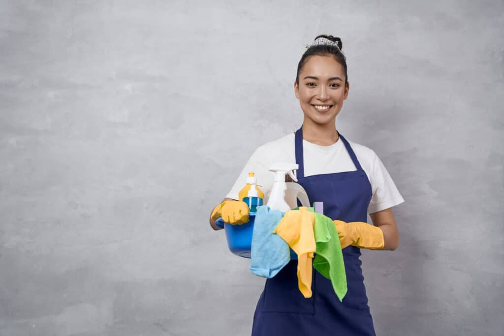 Femme de ménage souriante en tablier bleu et gants jaunes, tenant un seau avec produits d'entretien et lingettes colorées.