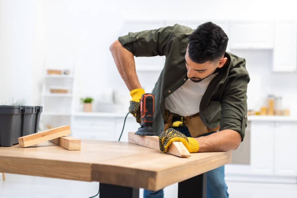 Homme barbu concentré, portant des gants de travail jaunes, ponce une planche de bois avec une ponceuse orbitale sur une table en bois clair.