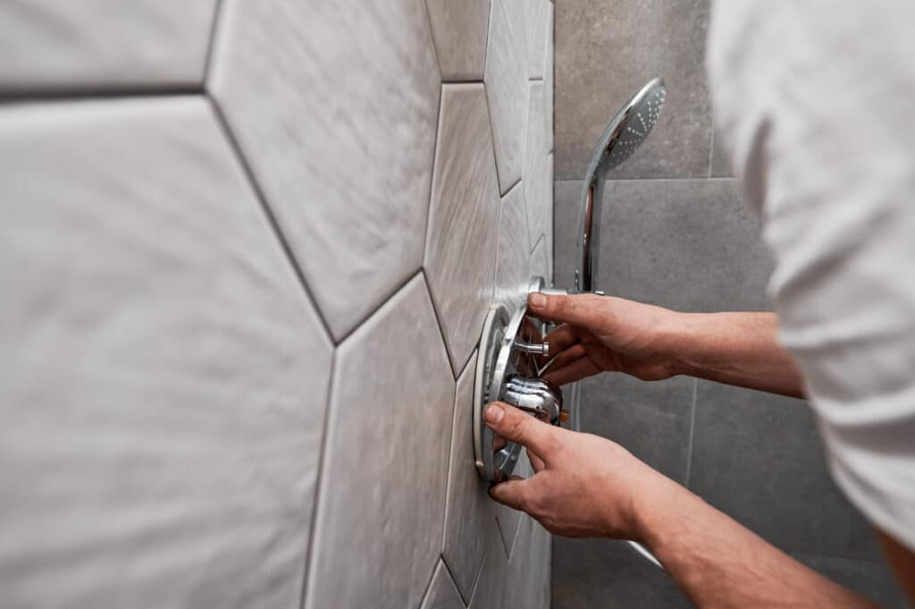 Mains de plombier installant ou ajustant la robinetterie chromée d'une douche moderne sur carrelage gris géométrique.