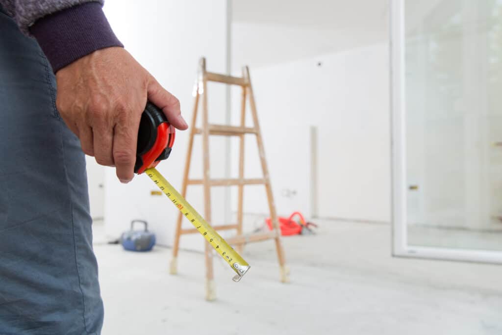 Gros plan sur une main tenant un mètre ruban jaune et rouge, affichant les nombres 8 à 15, dans une pièce en rénovation avec échelle.