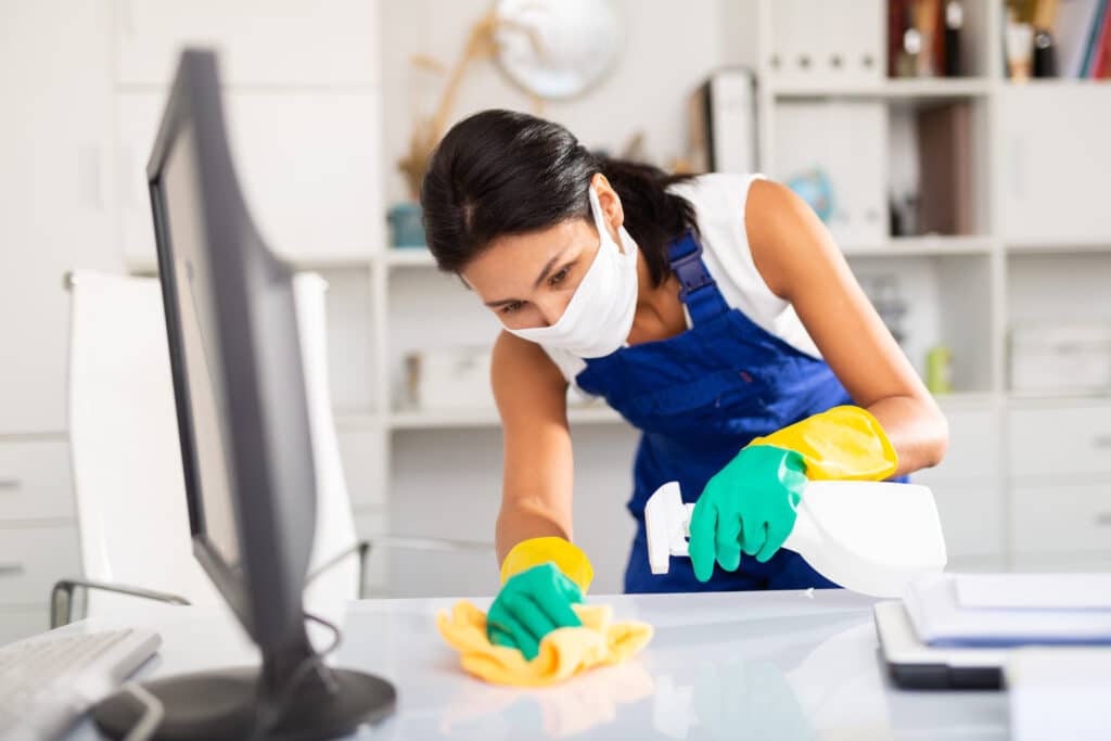 Femme en masque et uniforme bleu nettoie un bureau d'ordinateur blanc. Gants verts et jaunes, vaporisateur et chiffon.