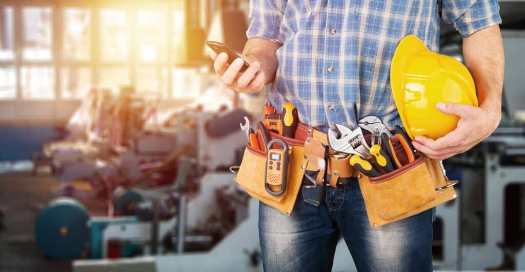 Ouvrier BTP en chemise à carreaux et ceinture à outils, tenant un smartphone et un casque jaune. Arrière-plan flou de chantier.