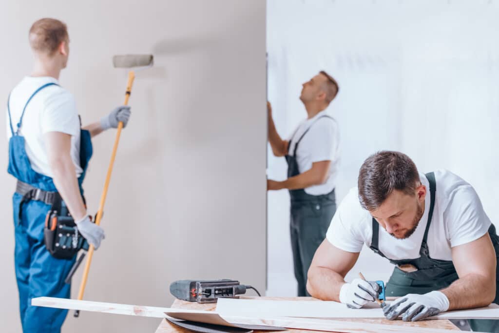 Trois ouvriers en rénovation: un peintre avec rouleau, un menuisier mesure du bois, et un ouvrier ajuste un mur. Travail de chantier.
